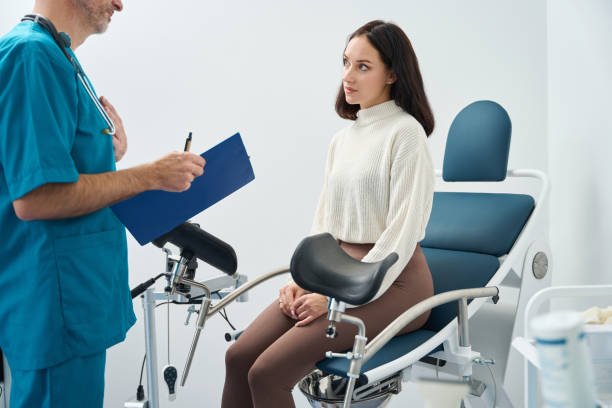 Женщина сидит в гинекологическом кресле для осмотра, в то время как медицинский работник разговаривает с ней.