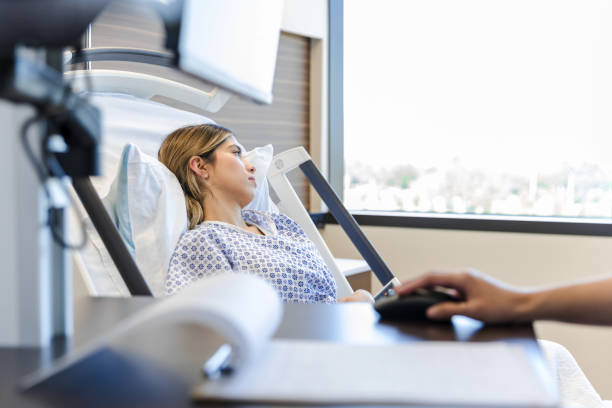 Женщина лежит на больничной койке у окна, а кто-то рядом работает за компьютером.